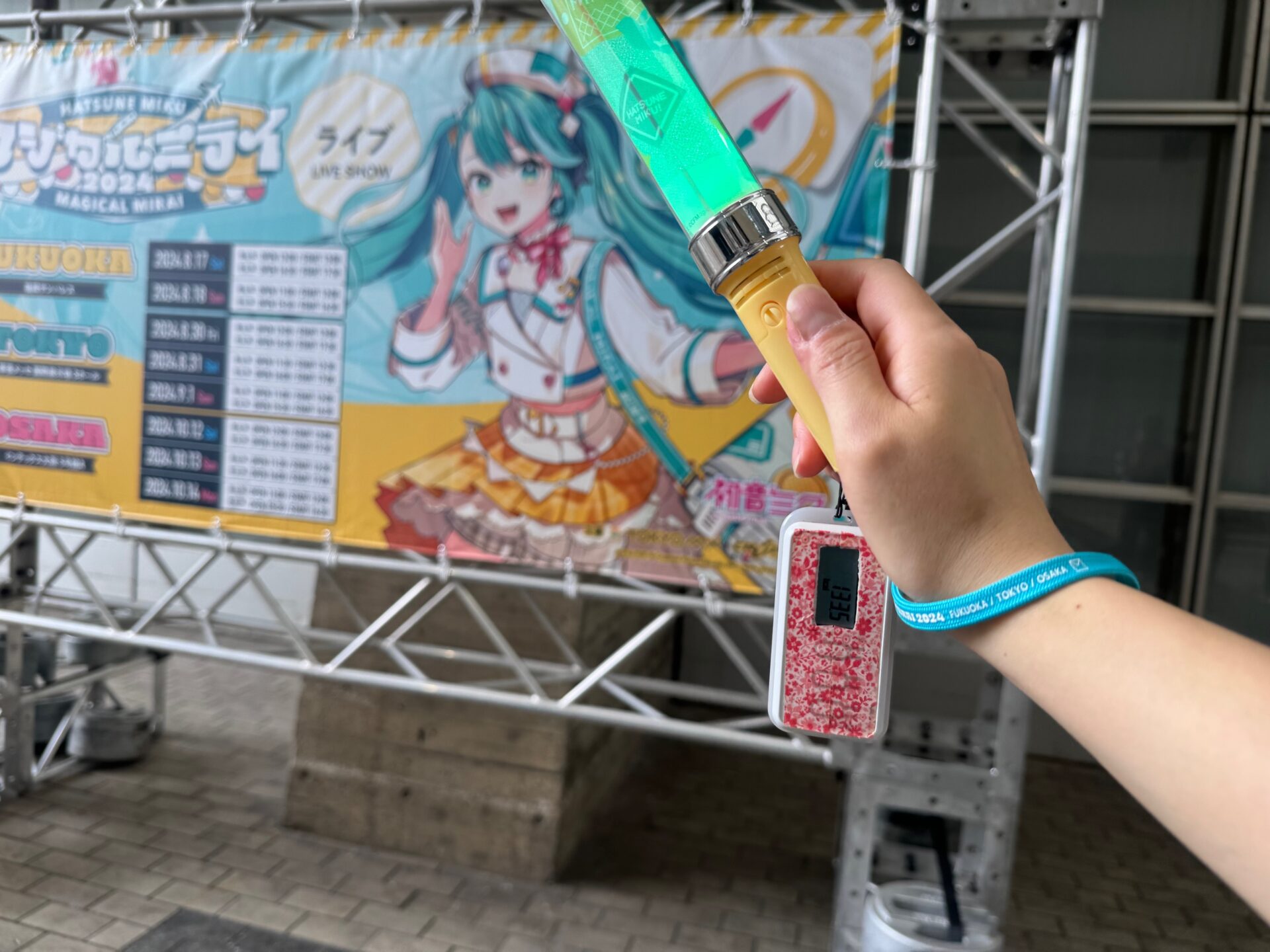 初音ミクのイベントの看板を背景に、ペンライトを手に持った写真。ペンライトにはタニタせいの活動量計がついています