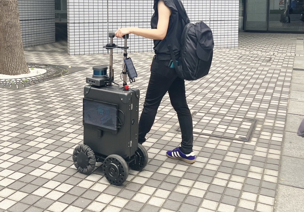 屋外用エーアイスーツケースに誘導されながら、未来館の外を歩く女性の写真