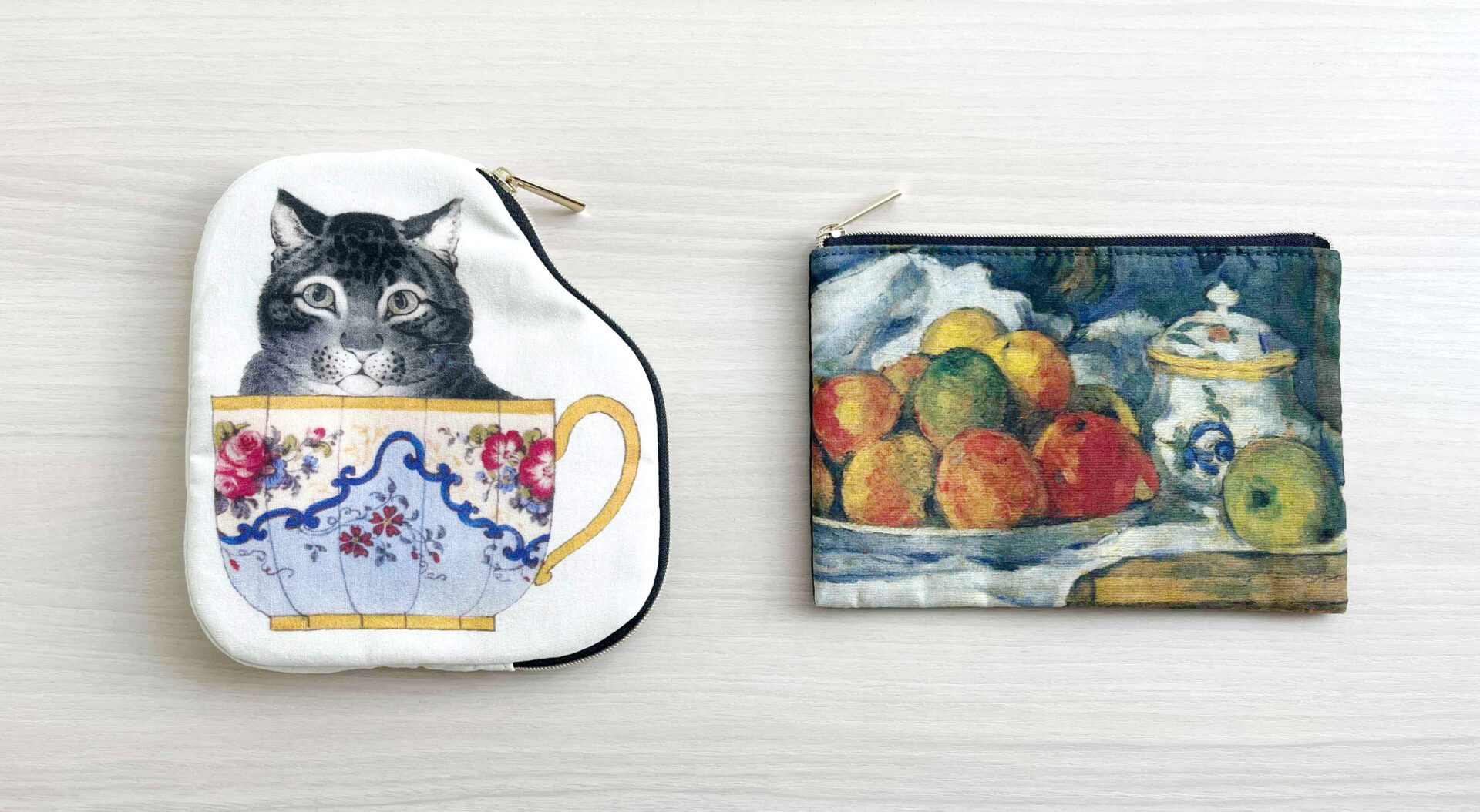 ティーカップにハイった猫のイラストが大きくえがかれたダイカットポーチとセザンヌのりんごプレートの絵画がえがかれたフラットポーチが並べておかれている写真