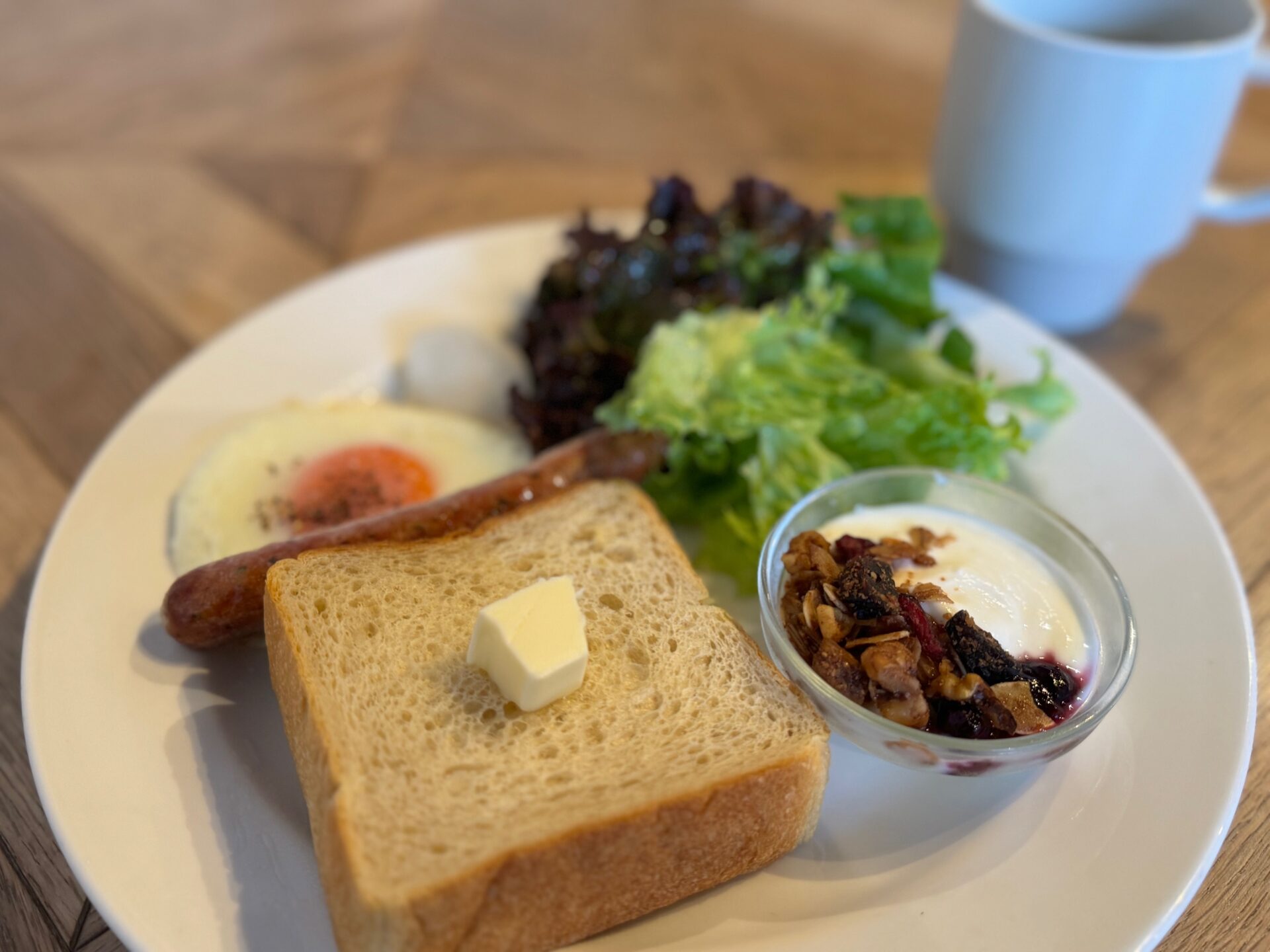 朝食プレートと、コーヒーの写真。プレートにはバターがのった食パン、目玉焼き、ソーセージ、葉野菜のサラダ、シリアルとジャムつきのヨーグルトがひとつのおさらにのっています